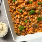 A white baking dish filled with baked pasta, meatballs, tomato sauce, and topped with fresh basil leaves brings comforting vibes, much like a classic Tuscan chicken pasta recipe. A small bowl of grated cheese sits nearby on a gray surface.