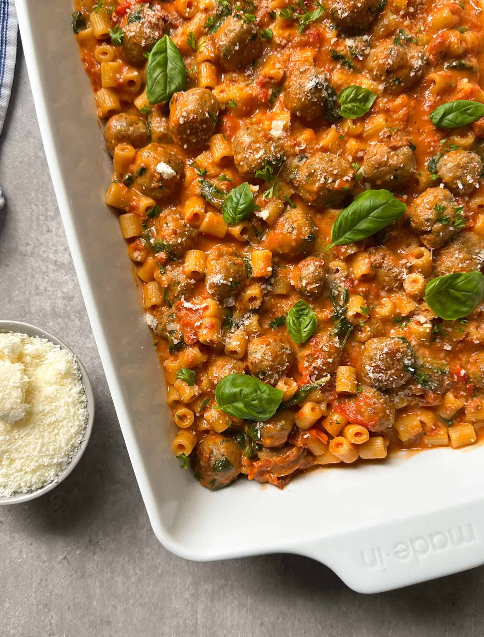A white baking dish filled with baked pasta, meatballs, tomato sauce, and topped with fresh basil leaves brings comforting vibes, much like a classic Tuscan chicken pasta recipe. A small bowl of grated cheese sits nearby on a gray surface.