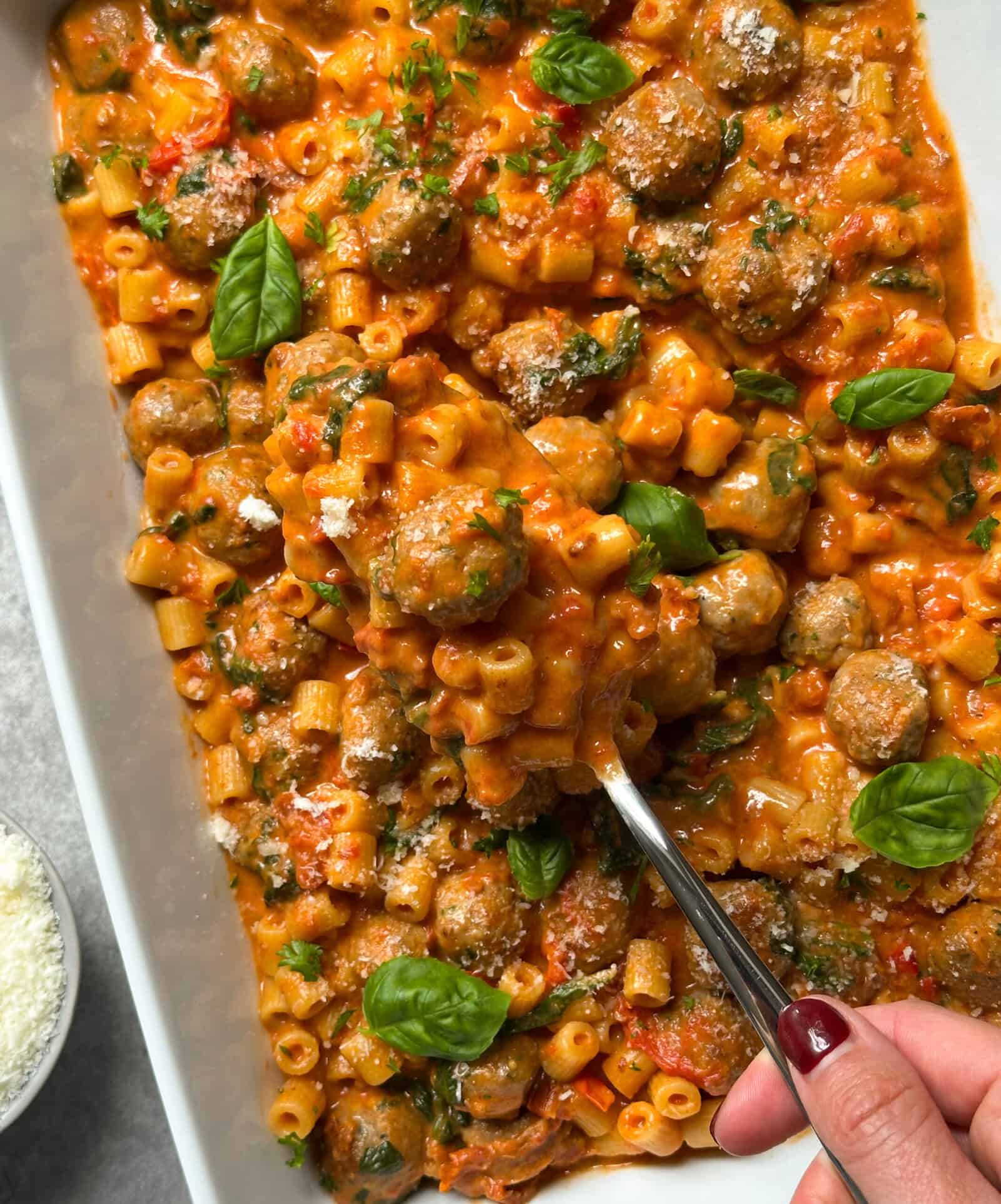 A hand holding a spoon serves a portion of creamy Tuscan chicken pasta with mini chicken meatballs, rich tomato sauce, and fresh basil from a casserole dish. The pasta is topped with grated cheese and garnished with basil leaves.