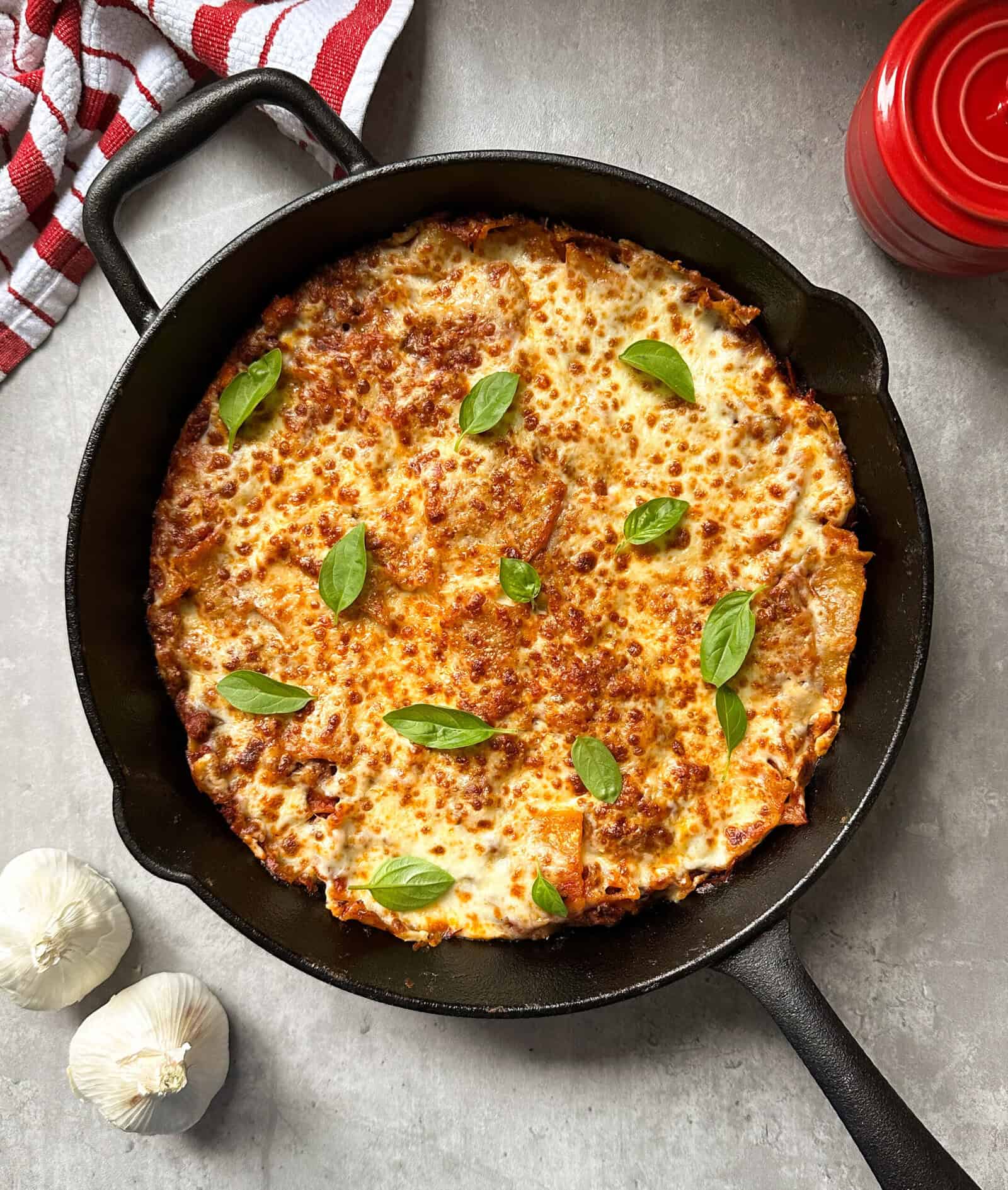 A skillet filled with easy lasagna, topped with melted cheese and fresh basil leaves, sits on a gray surface next to two bulbs of garlic, a red-and-white towel, and a red container—perfect for an easy lasagna recipe.