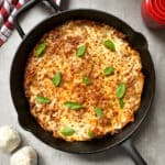 A skillet of easy lasagna topped with melted cheese and fresh basil sits on a gray surface, with a red-striped towel, a red container, and two garlic bulbs nearby.