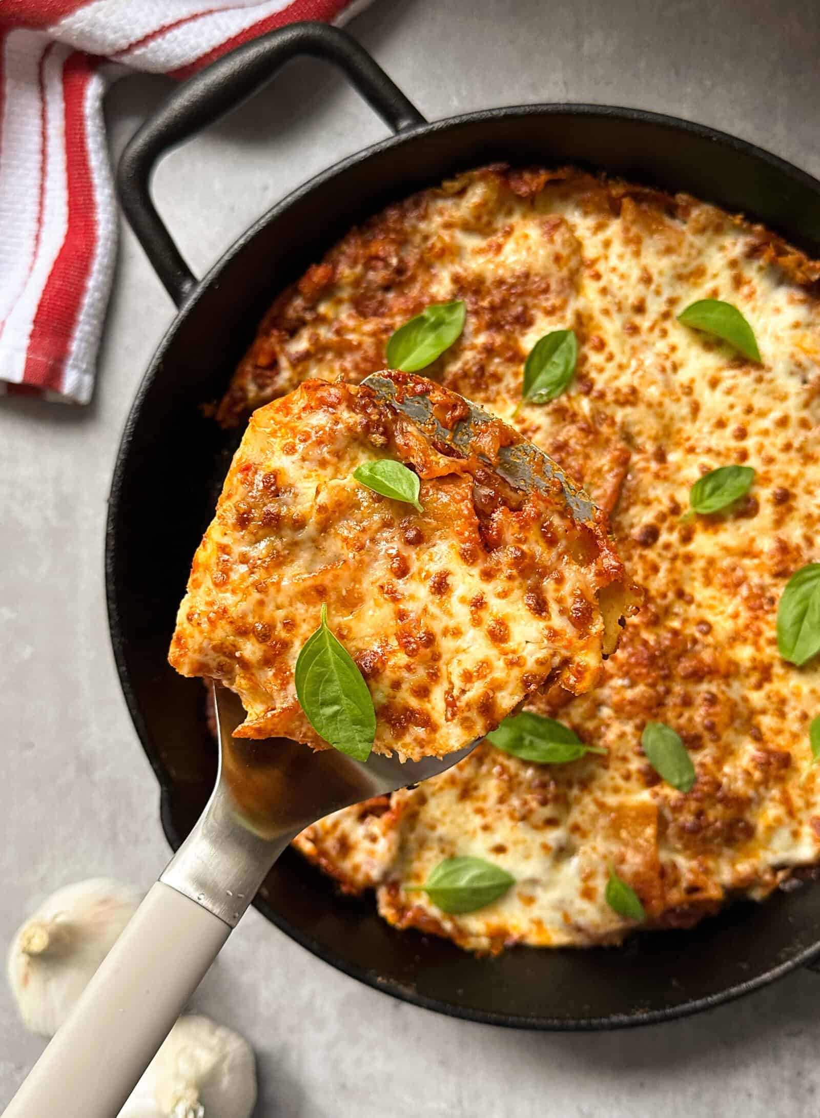 A cheesy, golden-brown slice of easy lasagna is lifted from a black skillet, topped with fresh basil leaves. The skillet lasagna sits on a gray surface beside a red-striped towel and garlic cloves.