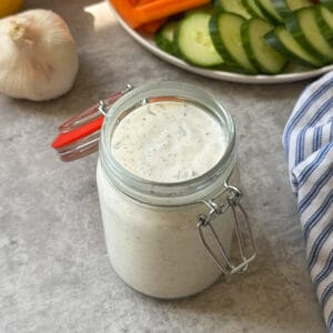 A glass jar of creamy homemade ranch dip sits on a gray surface next to a garlic bulb, with a plate of sliced cucumbers and carrot sticks in the background. A blue striped cloth is partially visible on the right.