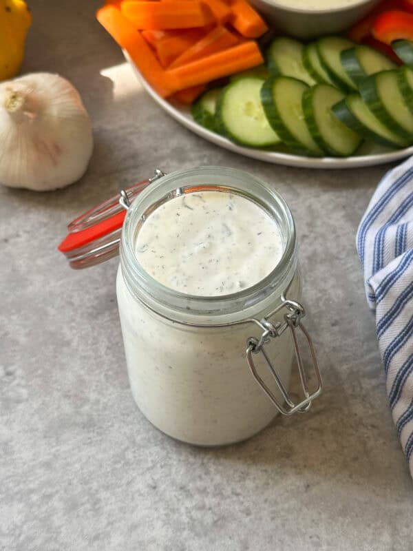 A glass jar of creamy homemade ranch dip sits on a gray surface next to a garlic bulb, with a plate of sliced cucumbers and carrot sticks in the background. A blue striped cloth is partially visible on the right.