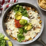 A skillet filled with a cheesy Mexican beef and rice skillet topped with sliced avocado, salsa, sour cream, cilantro, and a lime wedge. Tortilla chips and a red-striped towel are nearby on a gray surface.