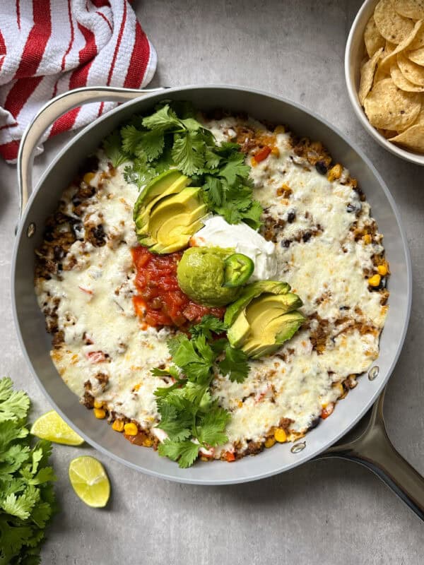 A Mexican beef and rice skillet filled with cheesy taco dip topped with avocado slices, salsa, sour cream, and fresh cilantro. Tortilla chips sit nearby on a gray surface, alongside lime wedges and a red striped towel for a vibrant touch.