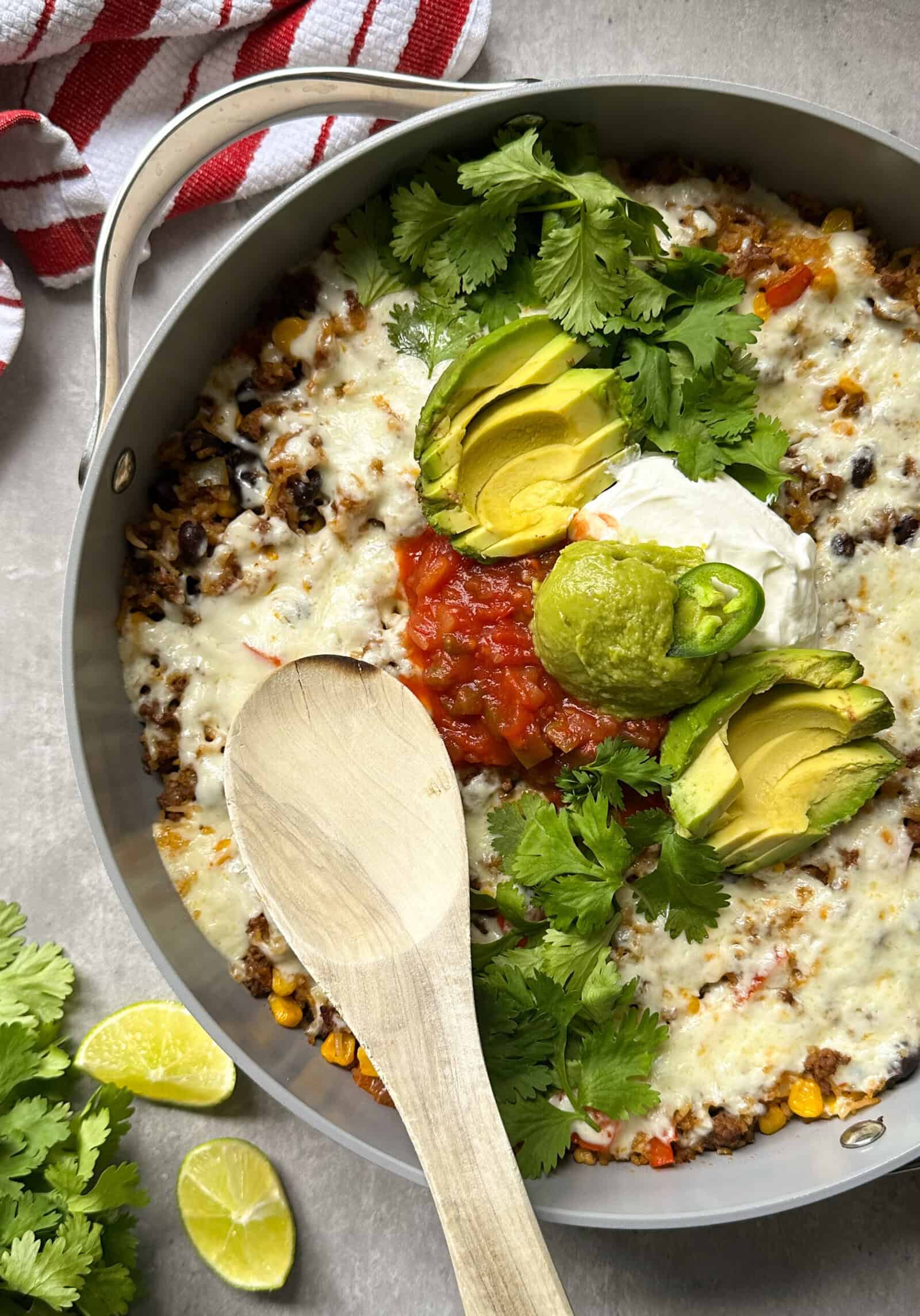 A cheesy Mexican skillet, topped with sliced avocado, salsa, sour cream, guacamole, and fresh cilantro, awaits with a wooden spoon inside. Lime wedges and extra cilantro sit nearby for this flavorful Mexican skillet recipe.