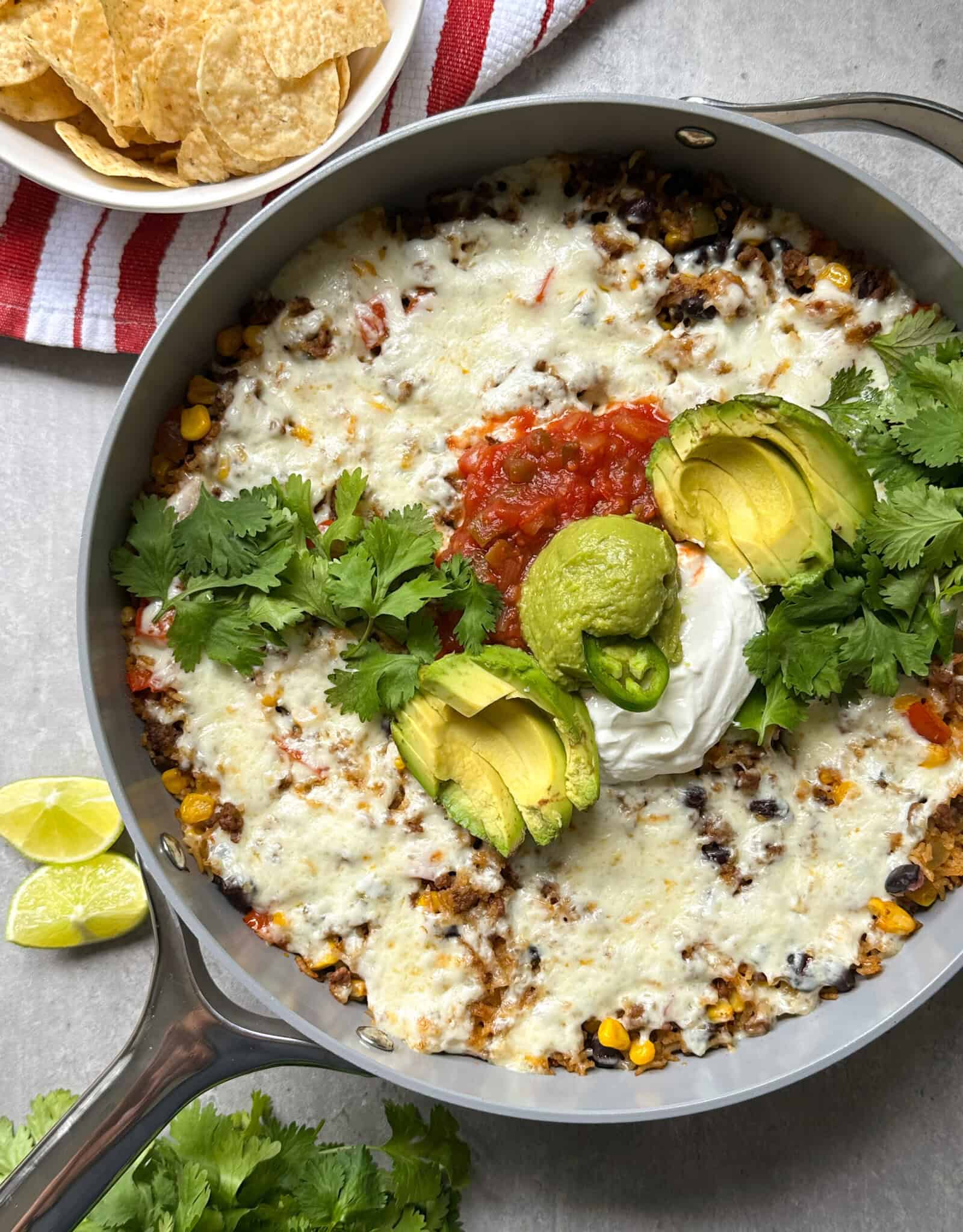 A skillet filled with a mouthwatering Mexican skillet recipe, topped with melted cheese, avocado slices, sour cream, salsa, guacamole, and fresh cilantro, served with lime wedges, tortilla chips, and a red-striped napkin.
