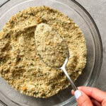 A hand holding a spoon scoops homemade bread crumbs from a clear glass bowl on a gray surface.