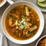 A bowl of chicken poblano soup recipe topped with crispy tortilla strips, cilantro, and a dollop of cream, served on a gray surface with lime wedges and extra tortilla strips on the side.