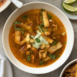 A bowl of chicken poblano soup recipe topped with crispy tortilla strips, cilantro, and a dollop of cream, served on a gray surface with lime wedges and extra tortilla strips on the side.