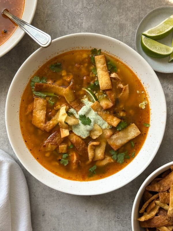 A bowl of chicken poblano soup recipe topped with crispy tortilla strips, cilantro, and a dollop of cream, served on a gray surface with lime wedges and extra tortilla strips on the side.