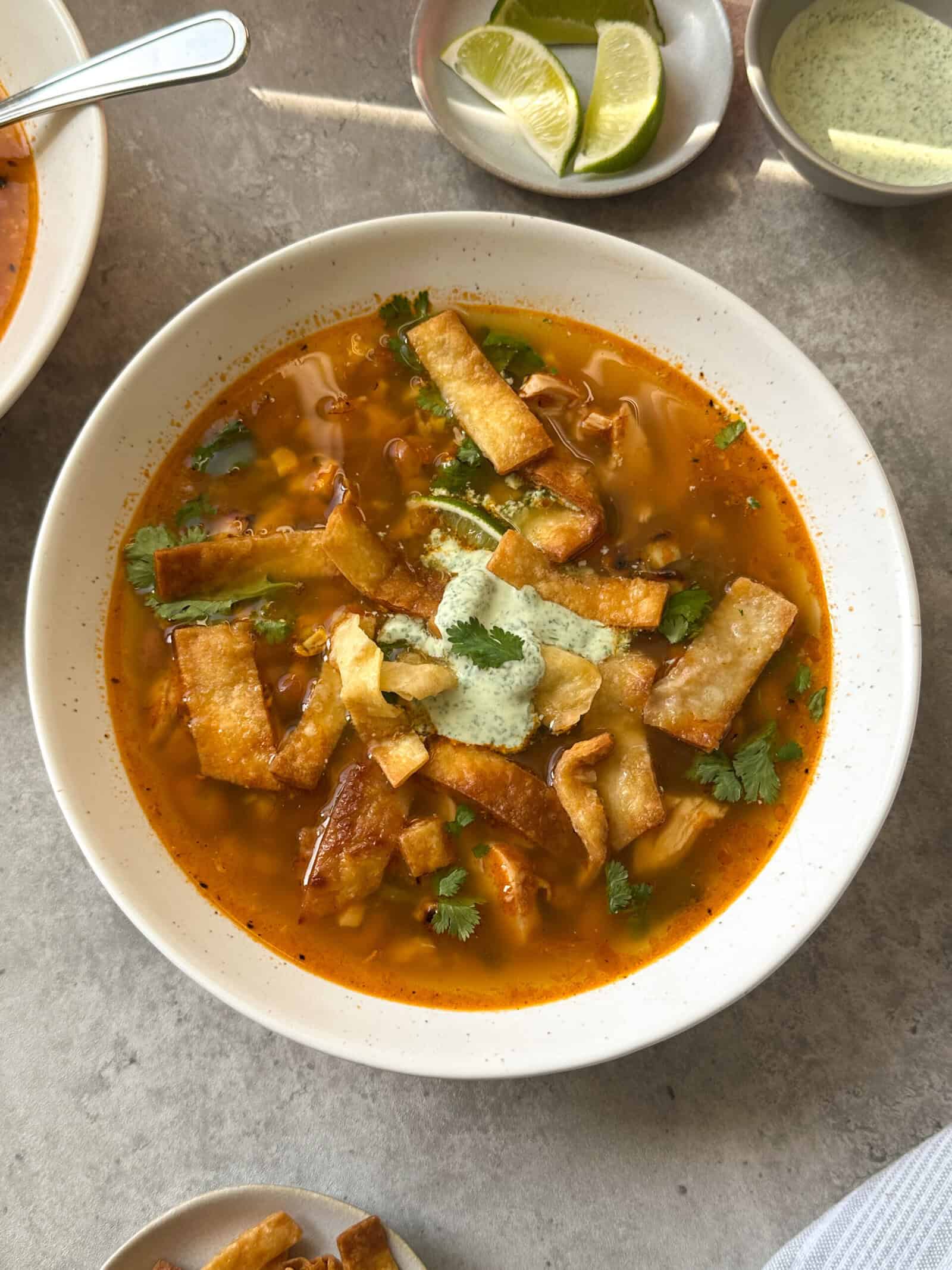A bowl of Chicken Poblano Soup garnished with crispy tortilla strips, fresh cilantro, and a dollop of green sauce. Slices of lime and a small bowl of green sauce are nearby on a light-colored surface.