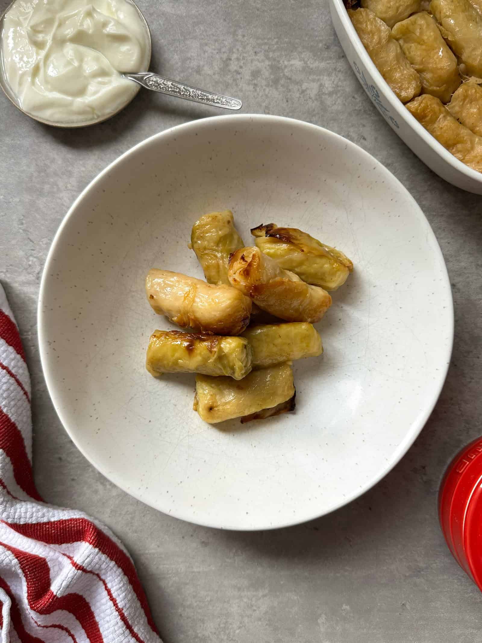 A white plate with several stuffed cabbage rolls on a gray surface. Nearby are a bowl of yogurt with a spoon, a red-and-white striped cloth, a red lid, and a dish of more cabbage rolls recipe favorites.