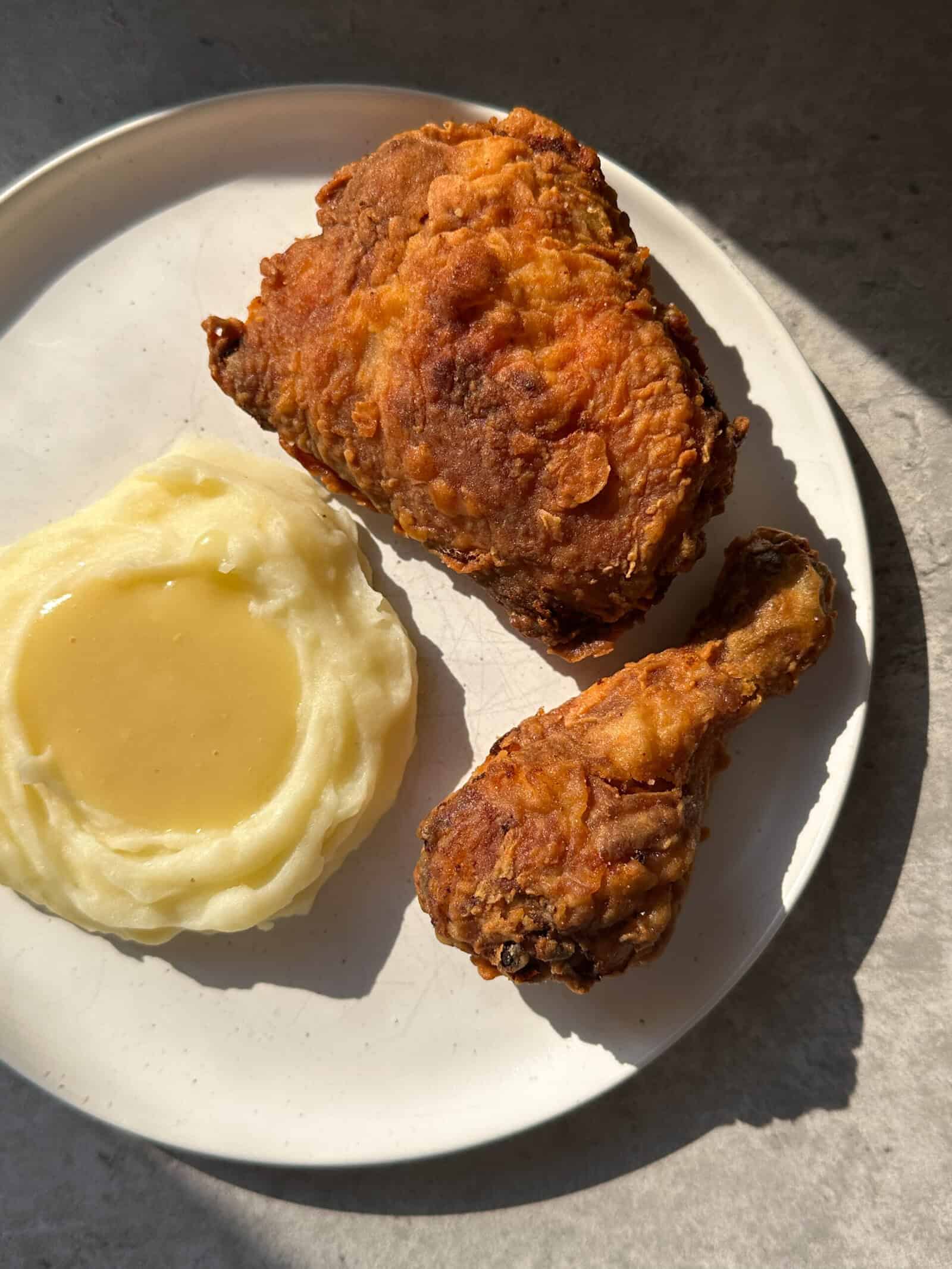 A plate of food with mashed potatoes and gravy, a piece of fried chicken, and a crispy fried chicken drumstick, all lit by natural sunlight—a perfect meal for anyone hunting for the best crispy fried chicken recipe.