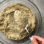 A hand with pale purple nail polish holds a spoon over a glass bowl filled with bread crumbs on a gray surface, perfect for your next homemade bread crumbs recipe.