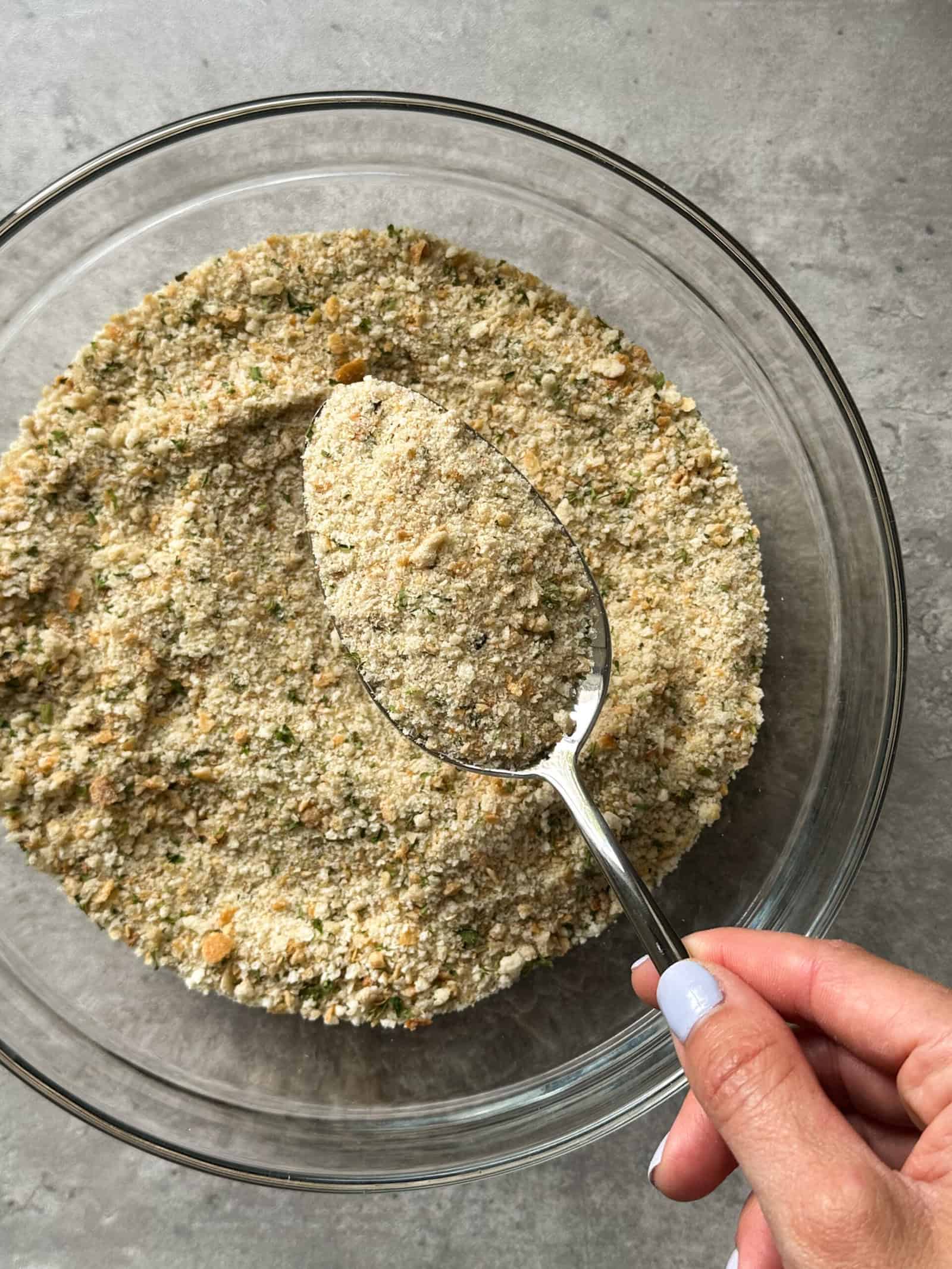 A hand with pale purple nail polish holds a spoon over a glass bowl filled with bread crumbs on a gray surface, perfect for your next homemade bread crumbs recipe.