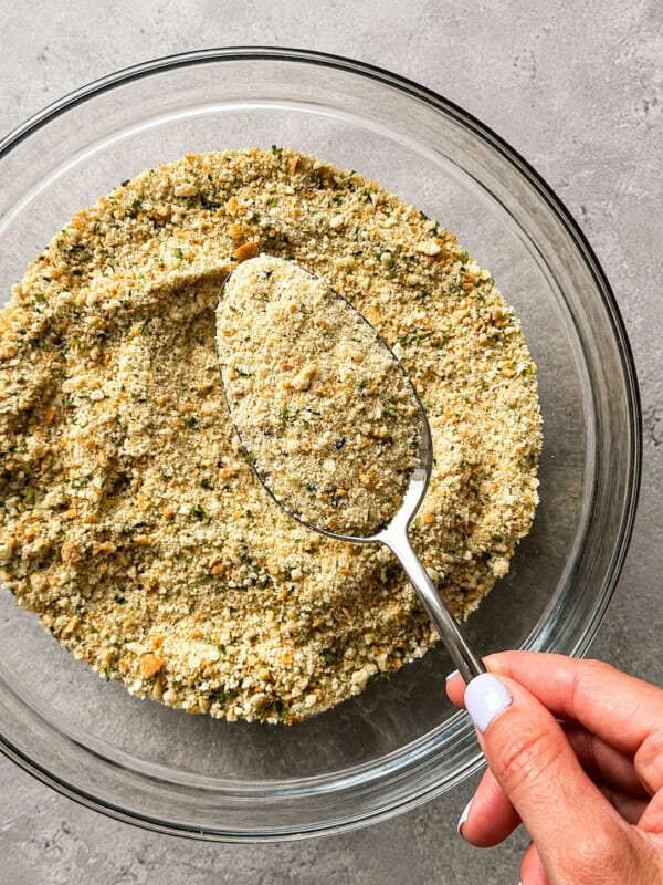 A hand holding a spoon scoops homemade bread crumbs from a clear glass bowl on a gray surface.