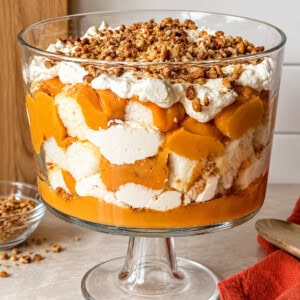 A layered pumpkin trifle in a glass pedestal bowl, topped with whipped cream and chopped nuts; this no-bake dessert is ready to serve.