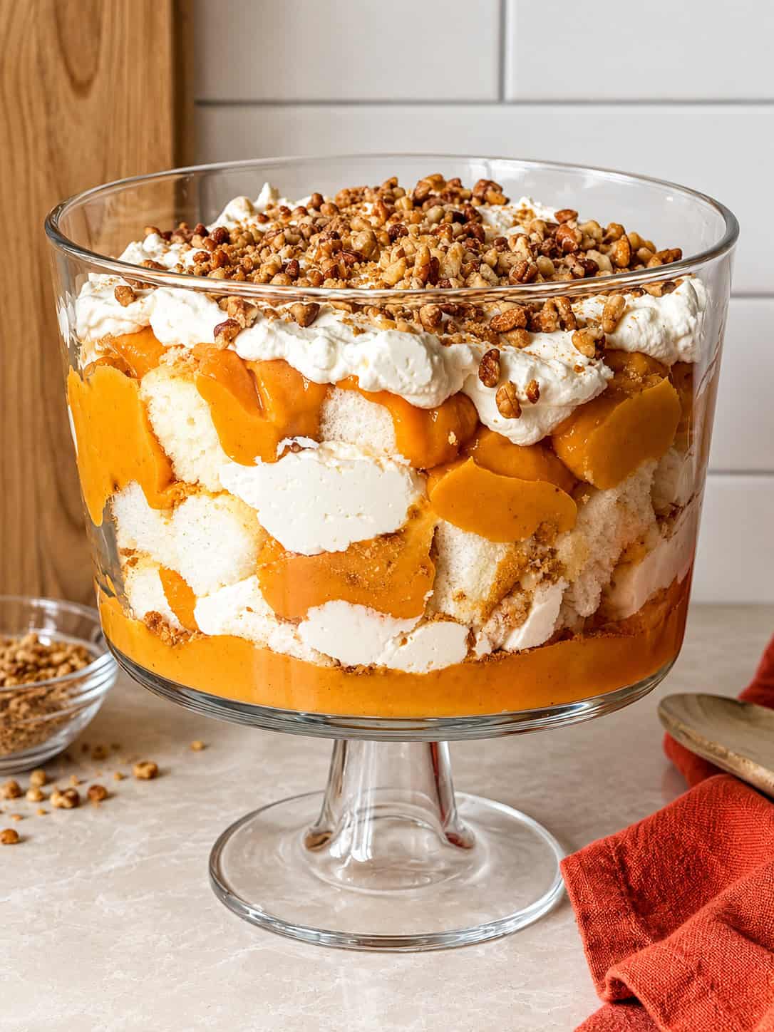 A layered pumpkin trifle in a glass pedestal bowl, topped with whipped cream and chopped nuts; this no-bake dessert is ready to serve.