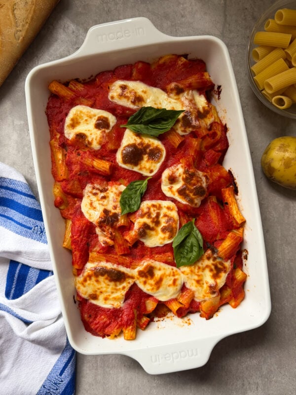 A white baking dish filled with Pasta e Patate Al Forno—baked rigatoni in tomato sauce with melted mozzarella and fresh basil. Nearby are a blue-striped towel, a baguette, uncooked pasta, and a potato.