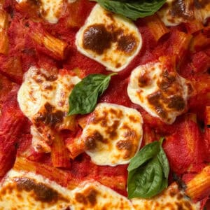 Close-up of baked ziti pasta topped with melted, browned mozzarella cheese, tomato sauce, and fresh basil leaves, similar to a Pasta e Patate Al Forno recipe.