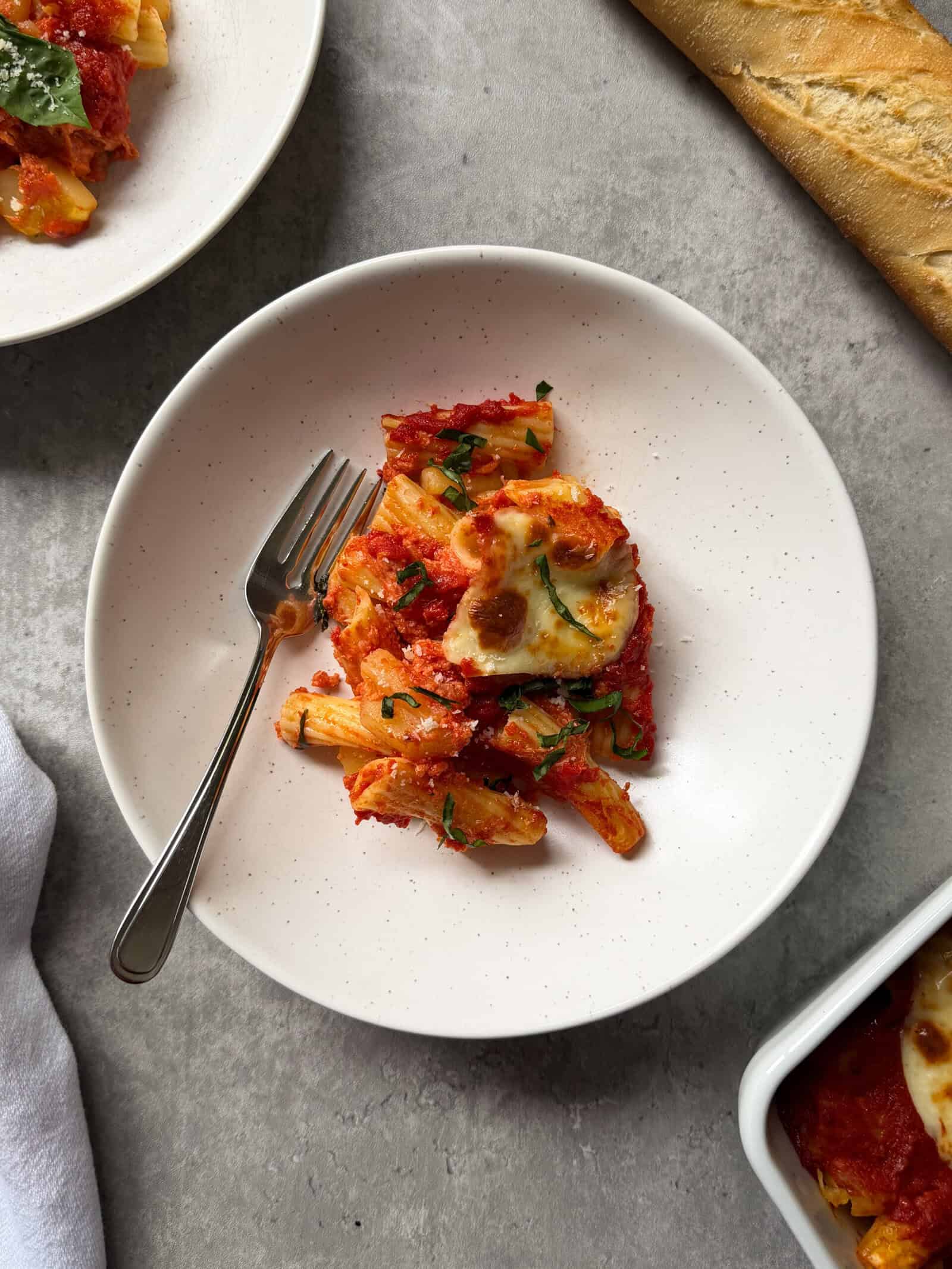 A white plate with baked rigatoni pasta in tomato sauce, inspired by Pasta e Patate Al Forno Recipe, is topped with melted cheese and fresh basil. It sits on a gray surface next to a fork, part of a baguette, and another plate of pasta.
