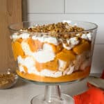 A glass trifle dish filled with a Pumpkin Trifle—layers of whipped cream, orange pudding or puree, cake pieces, and chopped nuts—sits on a light countertop with an orange cloth nearby. Perfect for fans of no bake desserts.