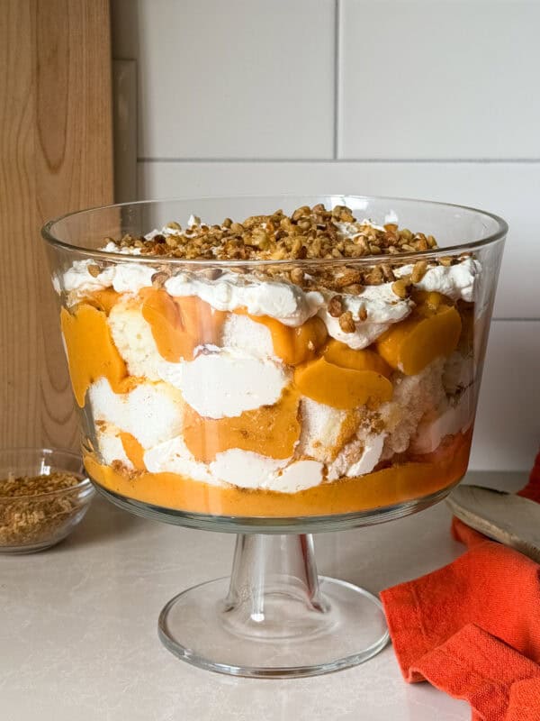 A glass trifle dish filled with a Pumpkin Trifle—layers of whipped cream, orange pudding or puree, cake pieces, and chopped nuts—sits on a light countertop with an orange cloth nearby. Perfect for fans of no bake desserts.