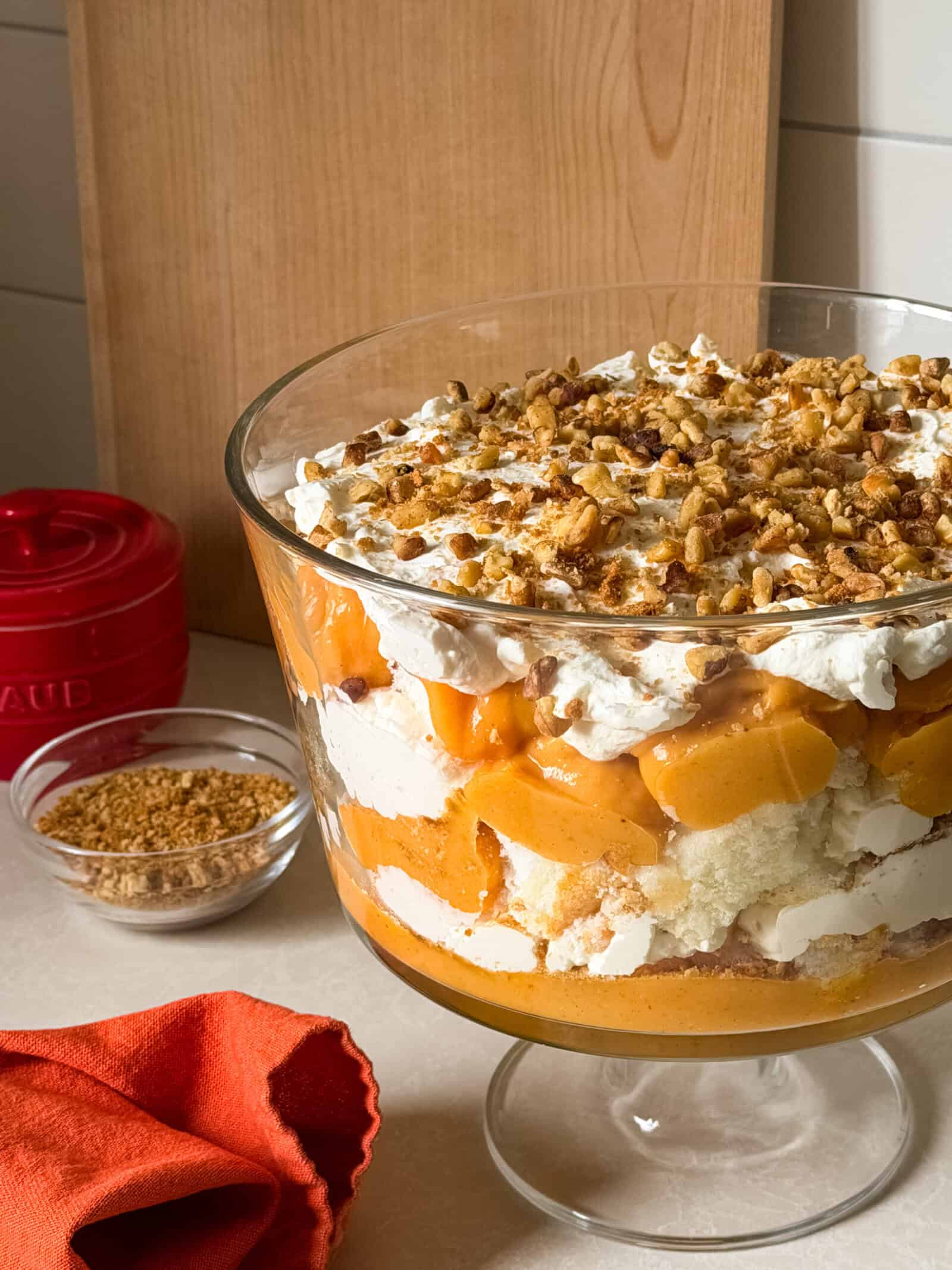 A glass trifle bowl filled with layers of whipped cream, sponge cake, and peaches, topped with chopped nuts—perfect for fans of No Bake Desserts. A small bowl of crumble and a red container sit in the background on a kitchen counter.