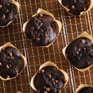 Double Chocolate Chip Muffins in brown paper liners are cooling on a wire rack. The muffins are topped with extra chocolate chips and arranged neatly on the wooden surface, perfect for any chocolate lover.
