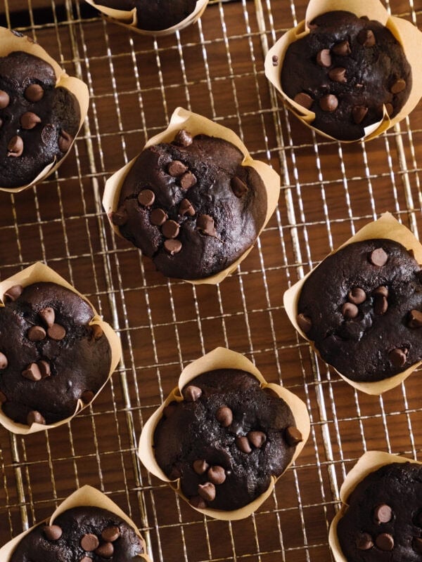 Double Chocolate Chip Muffins in brown paper liners are cooling on a wire rack. The muffins are topped with extra chocolate chips and arranged neatly on the wooden surface, perfect for any chocolate lover.