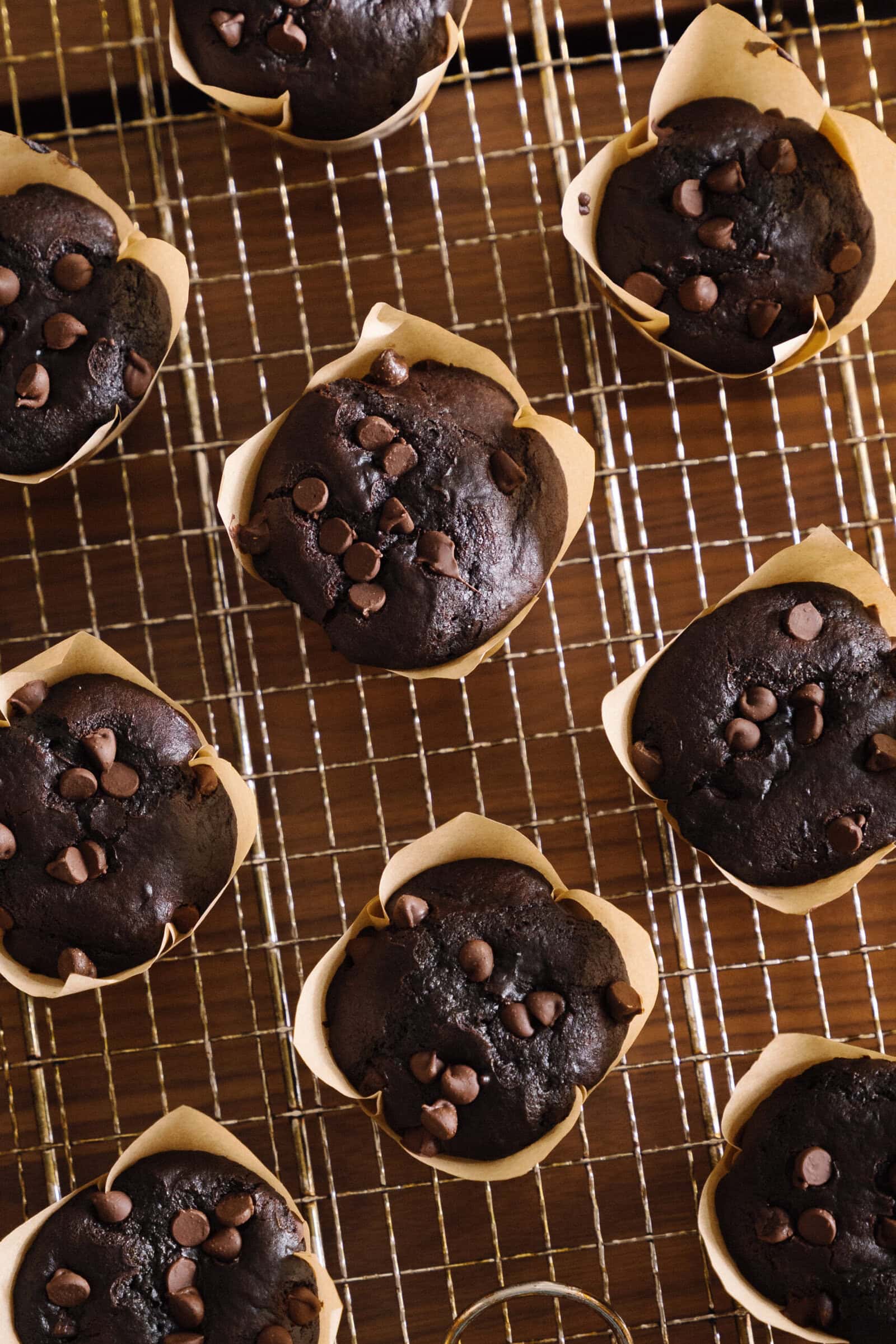 Double Chocolate Chip Muffins in brown paper liners are cooling on a wire rack. The muffins are topped with extra chocolate chips and arranged neatly on the wooden surface, perfect for any chocolate lover.