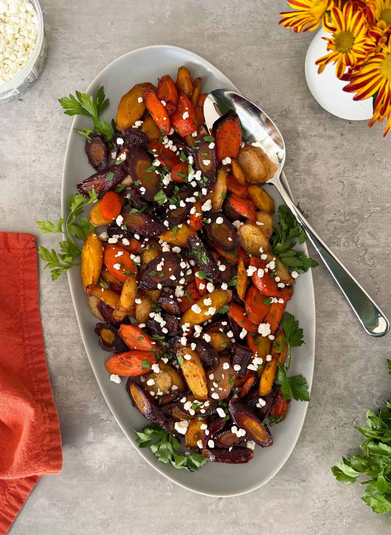 An oval platter of Honey Glazed Carrots, garnished with chopped herbs and crumbled white cheese, sits on a gray surface beside a spoon, red napkin, and a vase with orange-yellow flowers.