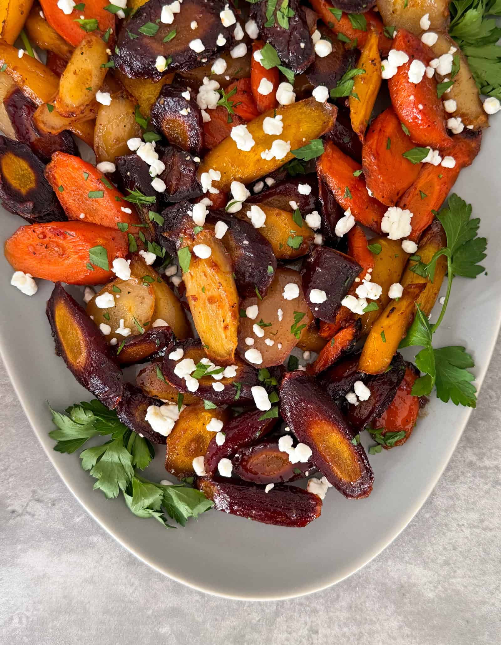 A platter of roasted multicolored honey carrots garnished with crumbled feta cheese and fresh parsley, served on a gray plate.