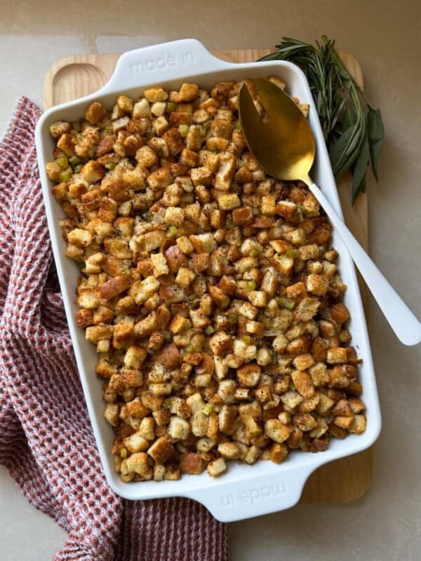 A white baking dish filled with golden brown homemade stuffing sits on a wooden board, next to a gold spoon and fresh herbs, with a red-checkered kitchen towel nearbyโperfect for anyone learning how to make stuffing from scratch.