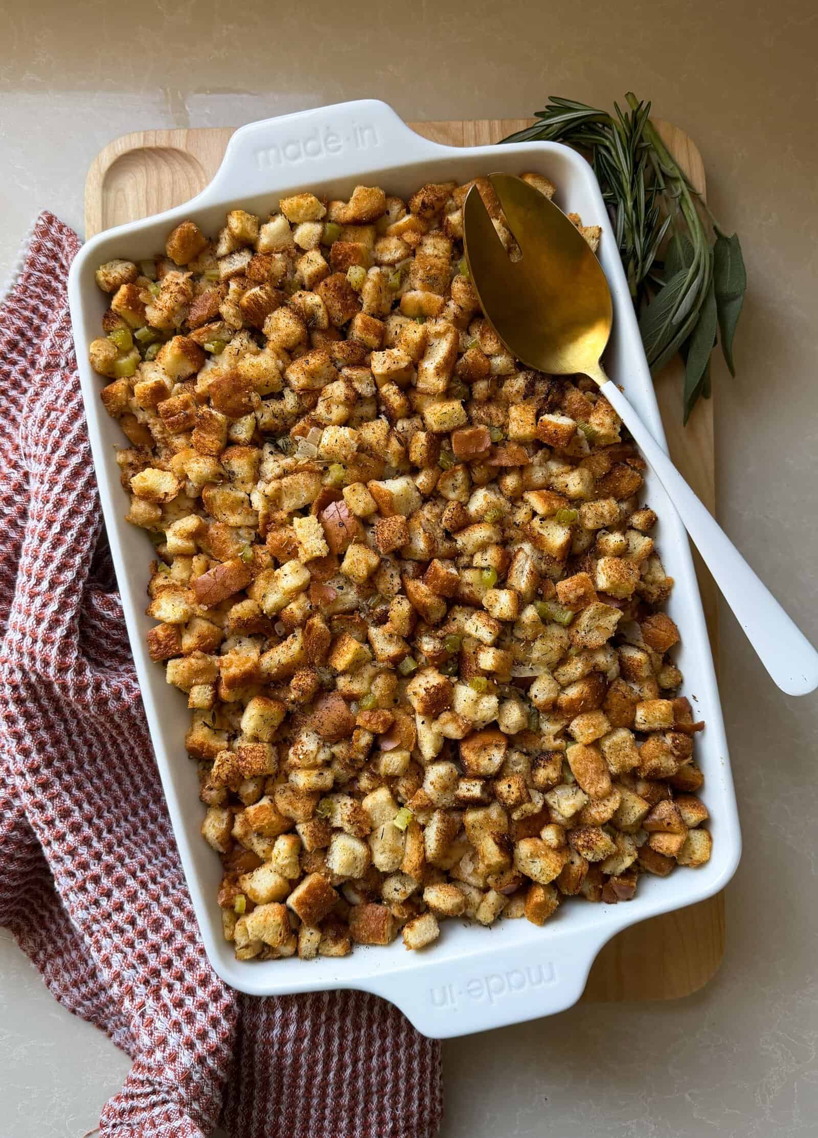 A white baking dish filled with golden brown homemade stuffing sits on a wooden board, next to a gold spoon and fresh herbs, with a red-checkered kitchen towel nearby—perfect for anyone learning how to make stuffing from scratch.