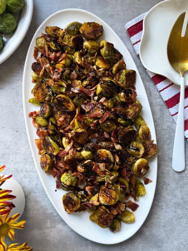A white oval platter filled with roasted Brussels sprouts mixed with crispy bacon pieces and garlic, placed on a gray surface next to a red-striped napkin—perfect for trying new Brussels Sprouts Recipes.