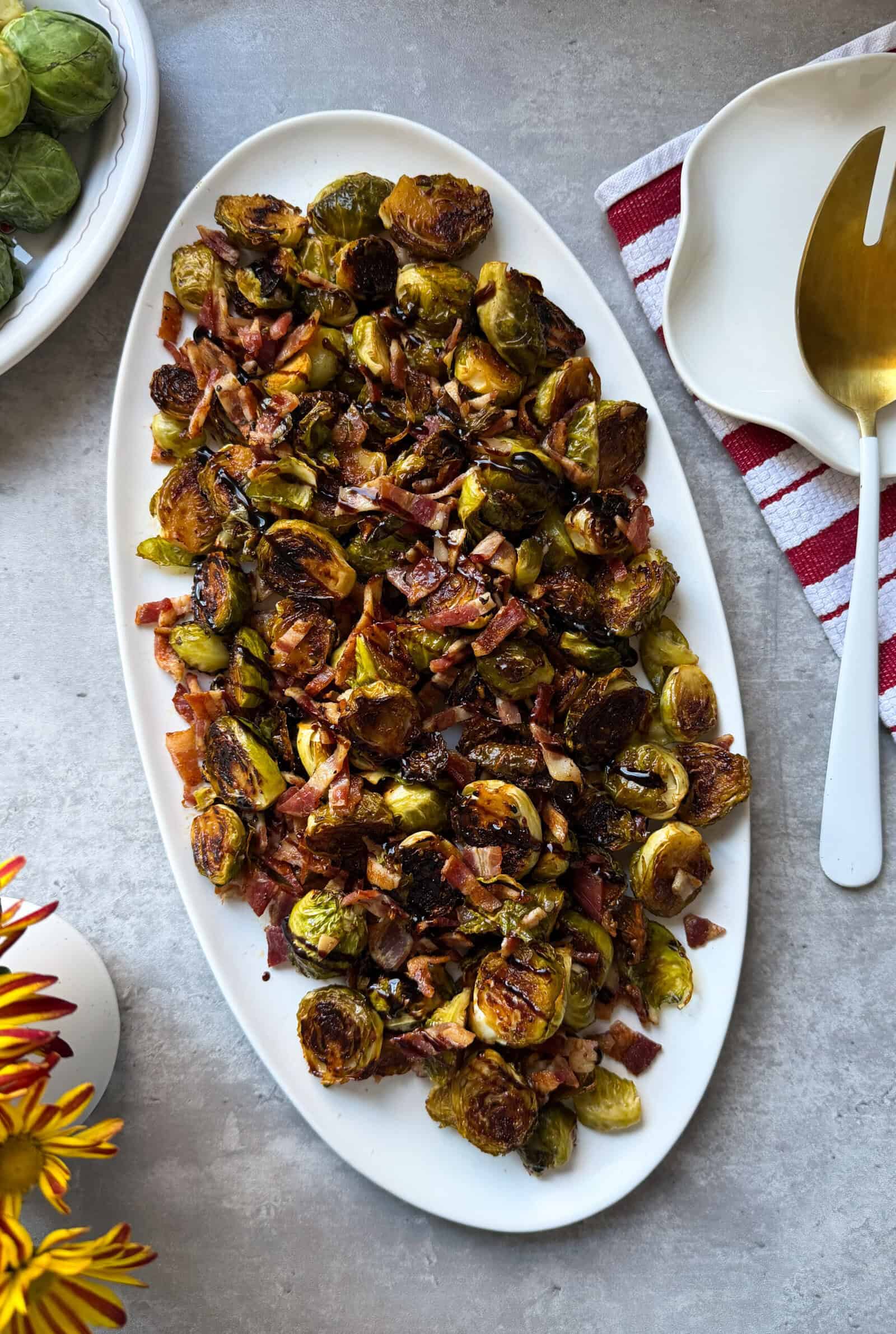 A white oval platter filled with roasted Brussels sprouts mixed with crispy bacon pieces and garlic, placed on a gray surface next to a red-striped napkin—perfect for trying new Brussels Sprouts Recipes.