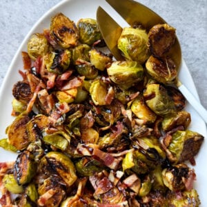 A white platter filled with Roasted Brussels Sprouts topped with chopped bacon and a dark balsamic glaze, alongside gold serving utensils on a gray background. This Roasted Brussels Sprouts recipe is perfect for any occasion.