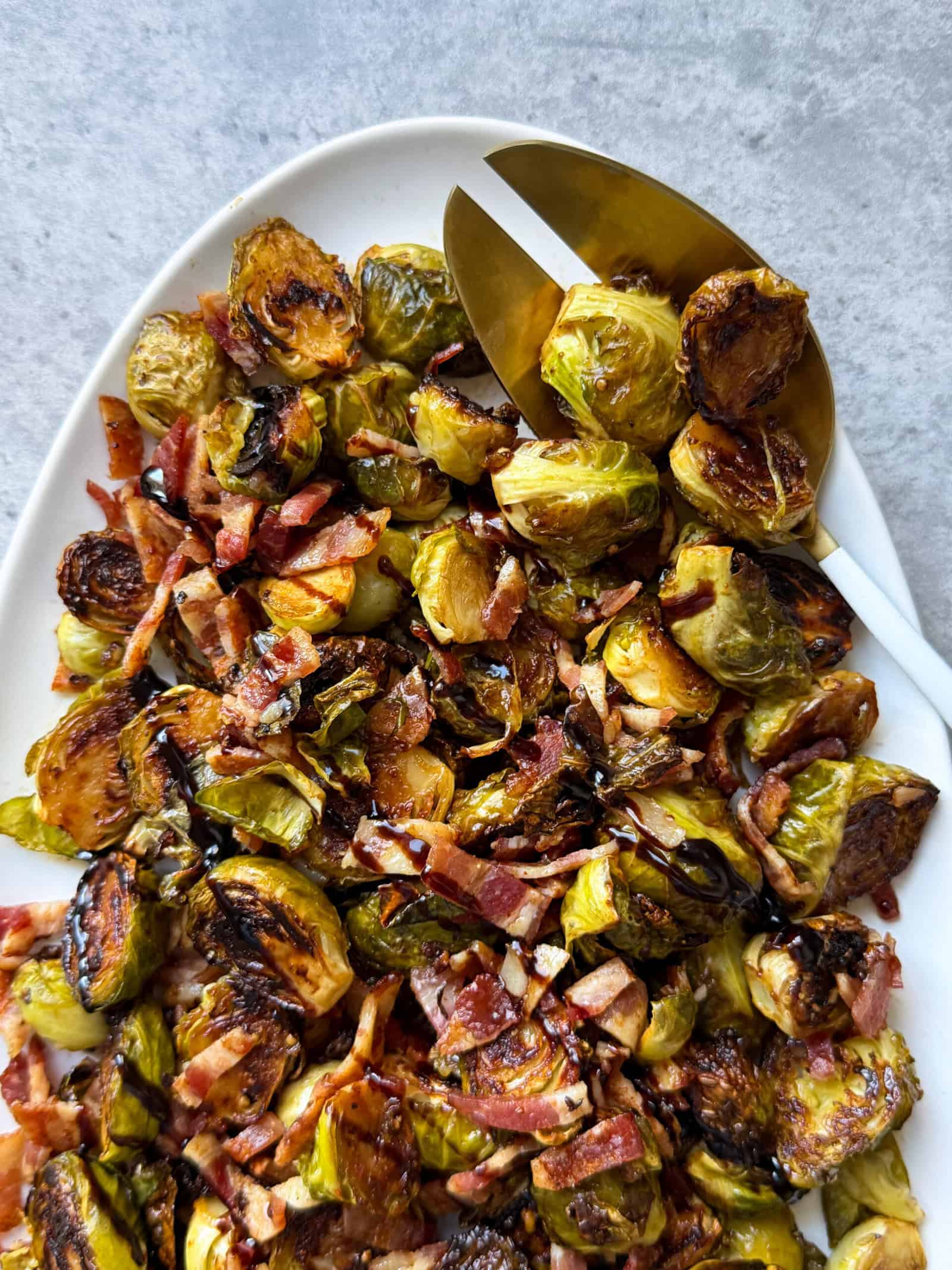 A white platter filled with Roasted Brussels Sprouts topped with chopped bacon and a dark balsamic glaze, alongside gold serving utensils on a gray background. This Roasted Brussels Sprouts recipe is perfect for any occasion.