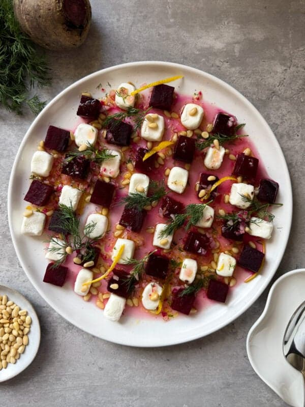 A white plate with cubes of beet, dollops of creamy cheese, pine nuts, fresh dill, and orange zest on a gray surface—an artful Beet Salad with Goat Cheese. Pine nuts and a beetroot sit nearby as garnish.