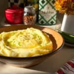 A bowl of creamy mashed potatoes, made from the perfect Creamy Mashed Potatoes Recipe, is garnished with chopped chives and sits on a countertop among kitchen items in warm sunlight.