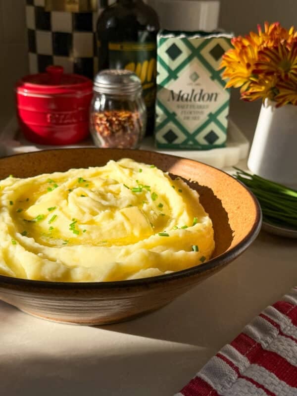 A bowl of creamy mashed potatoes, made from the perfect Creamy Mashed Potatoes Recipe, is garnished with chopped chives and sits on a countertop among kitchen items in warm sunlight.