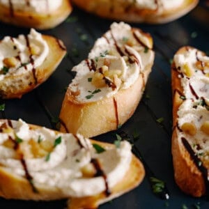 Sliced baguette topped with creamy cheese, chopped nuts, fresh herbs, and a drizzle of balsamic glaze, arranged on a dark plate.