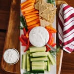 A rectangular platter featuring carrot sticks, cucumber sticks, red bell pepper strips, crackers, and a bowl of creamy dill dip. Try this easy veggie dip for your next gathering! A red and white striped napkin sits beside the platter on a wooden surface.