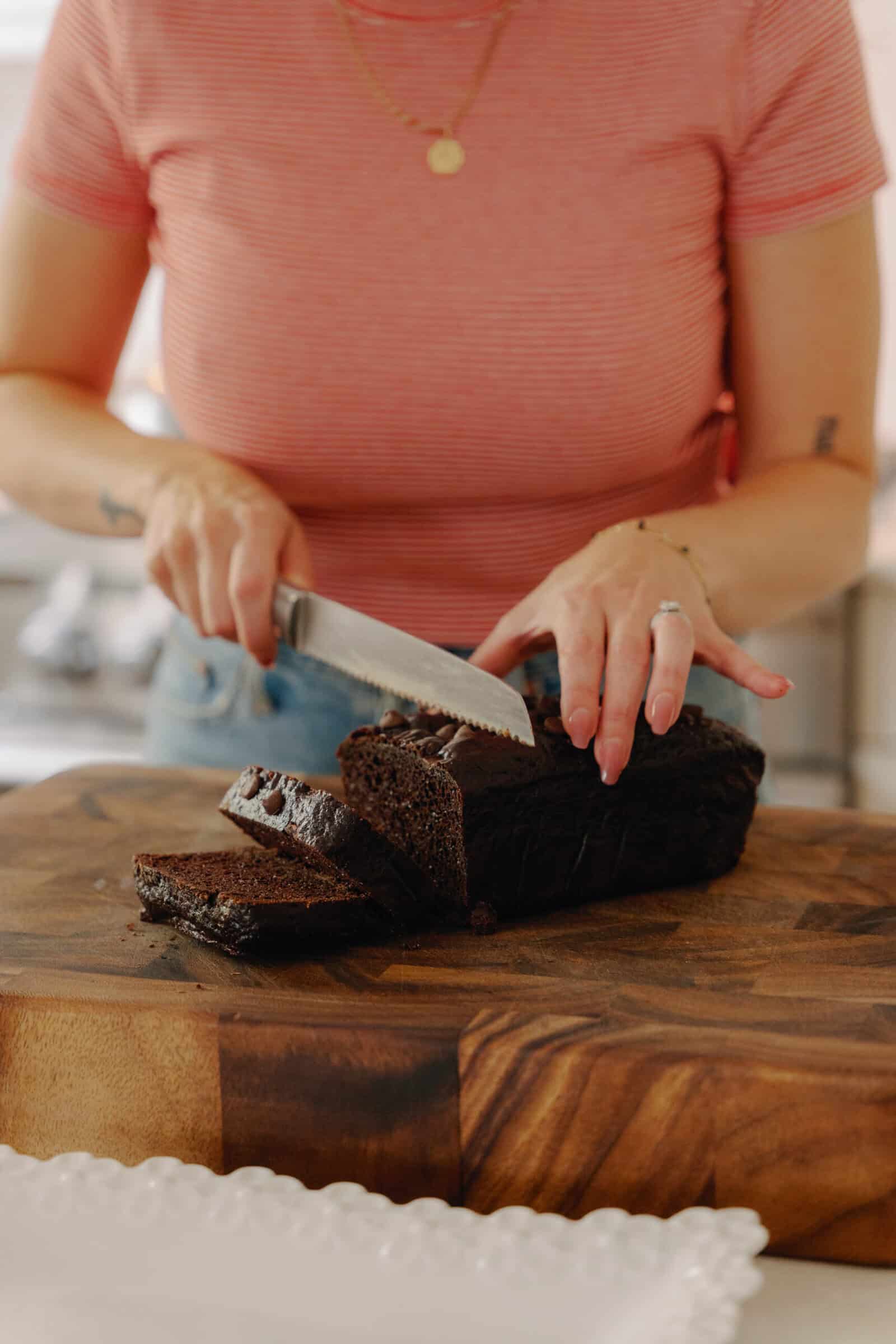 A person in a red striped shirt slices a chocolate loaf on a wooden cutting board in a kitchen. The focus is on their hands and the deliciously rich bread, perfect for trying out your favorite Chocolate Loaf Cake Recipe.