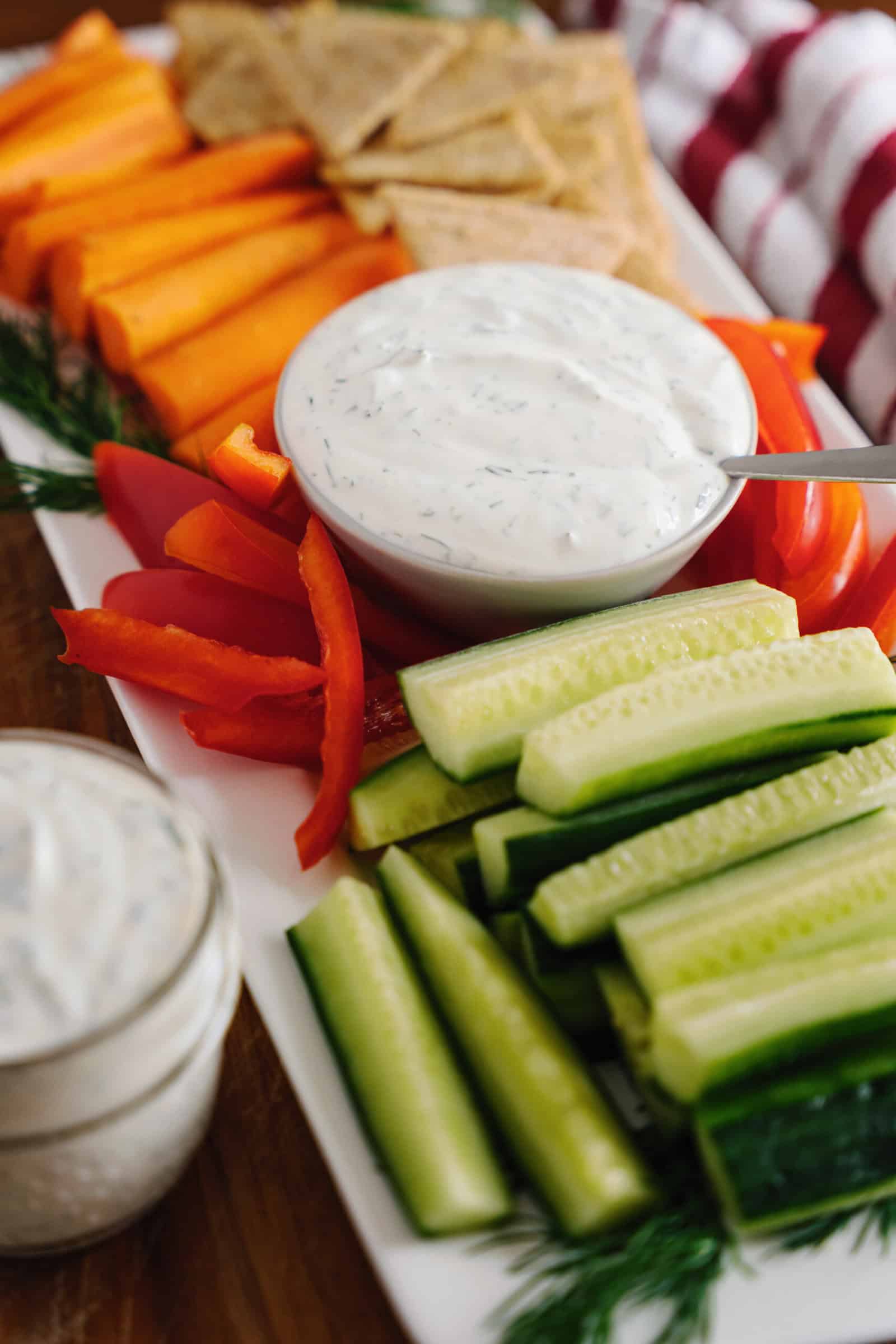 A platter with sliced cucumbers, red bell peppers, carrot sticks, pita chips, and a bowl of creamy dill dip garnished with fresh dill. Another small jar of veggie dip is nearby.