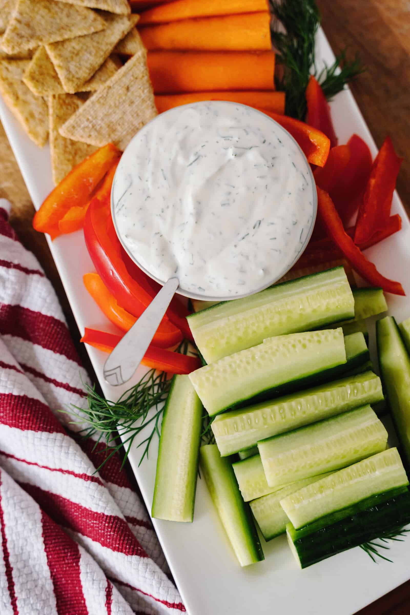A platter of fresh vegetables and crackers surrounds a bowl of creamy dill dip, perfect for any gathering. This veggie dip is served with cucumber sticks, red bell pepper slices, and carrots, alongside a striped red and white napkin.