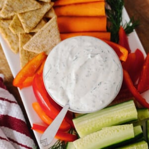 A platter with fresh vegetables, including cucumber sticks, carrot sticks, and red bell pepper strips, along with crackers and a bowl of creamy ranch dip, garnished with dill. A red and white striped towel is beside the platter.
